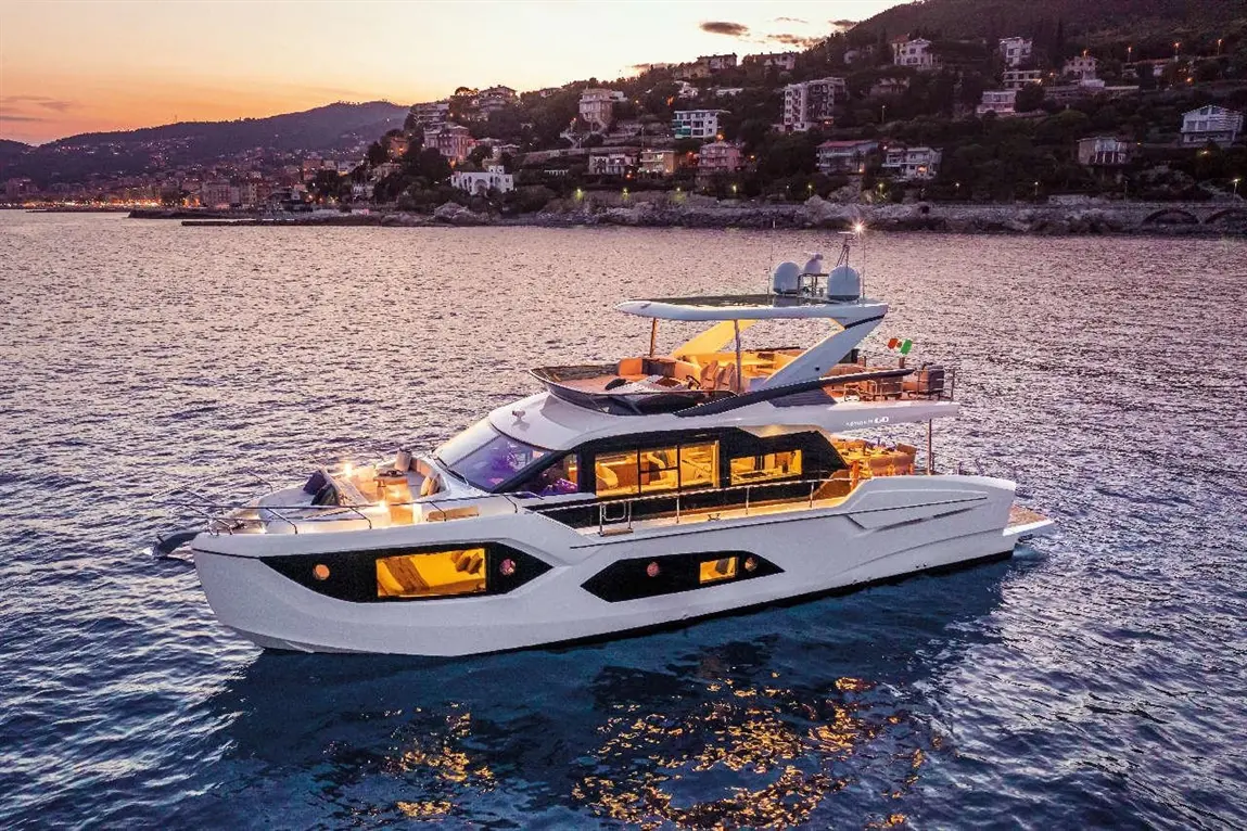 Luxury yacht anchored in calm waters at sunset, showcasing modern design and warm lighting with hills and houses in the background.